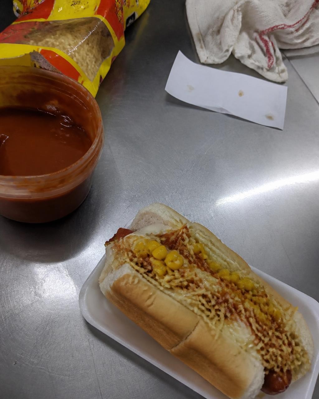 Foto do cachorro-quente na bancada de inox, com a bagunça do serviço atrás